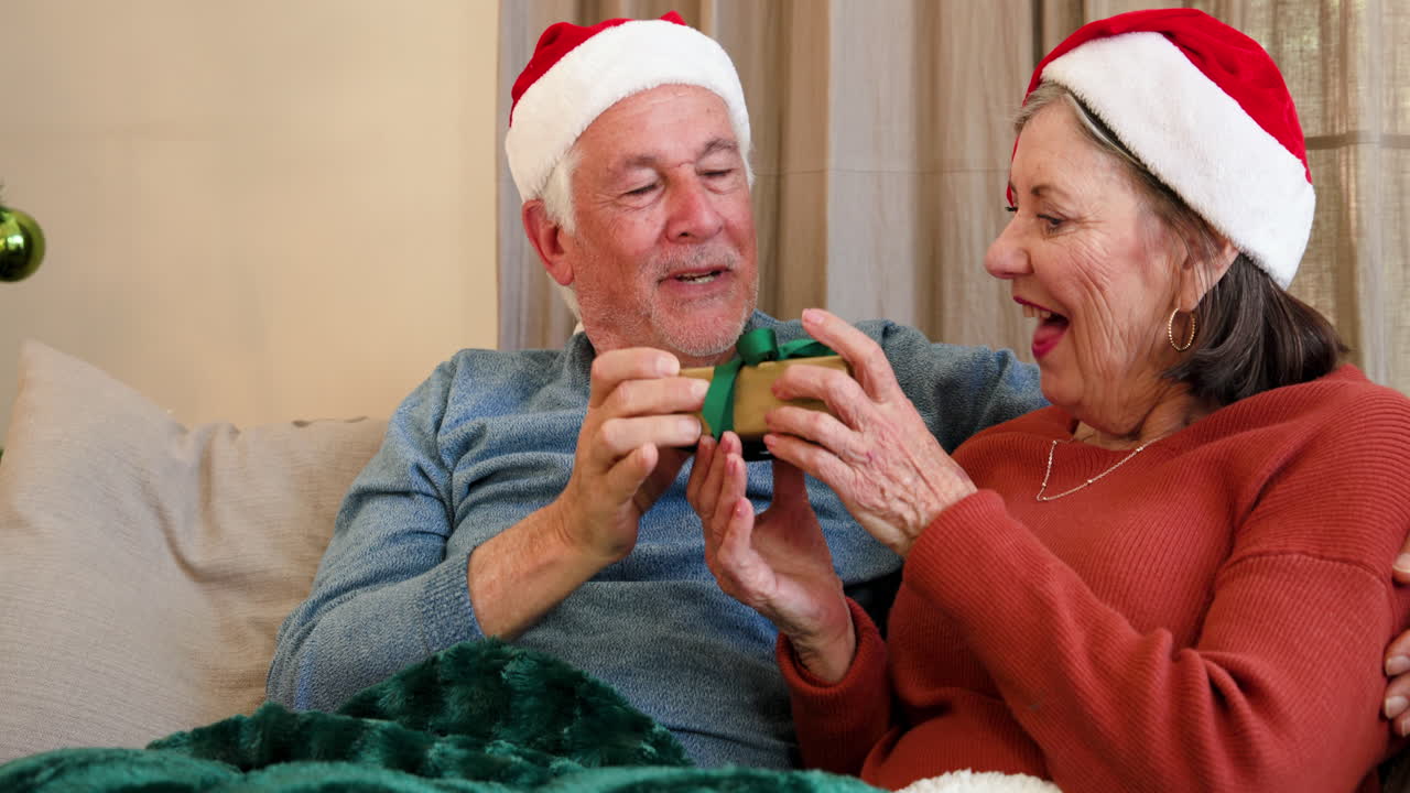 Senior couple wearing Santa hats enjoying Christmas gift exchange at home