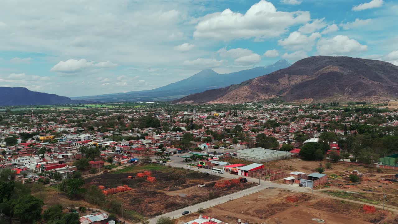 Outskirts of Tuxpan with residential area, open lots, Colima Volcano and Nevado de Colima in the background, aerial pan left