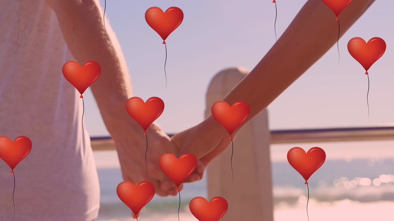 múltiples globos de corazón flotando contra la sección media de la pareja de la mano en la playa