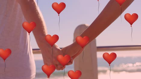 multiple heart balloons floating against mid section of couple holding hands at beach