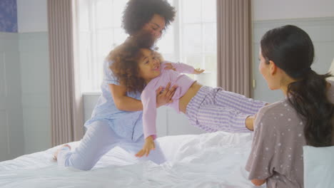 family with two mums wearing pyjamas playing on bed at home with daughter