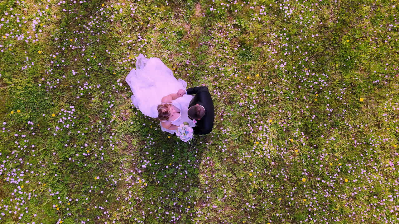 Bride in flowing gown and groom in formal suit share a dance on a sunlit meadow, viewed directly from above with shadows stretching across scattered wildflowers