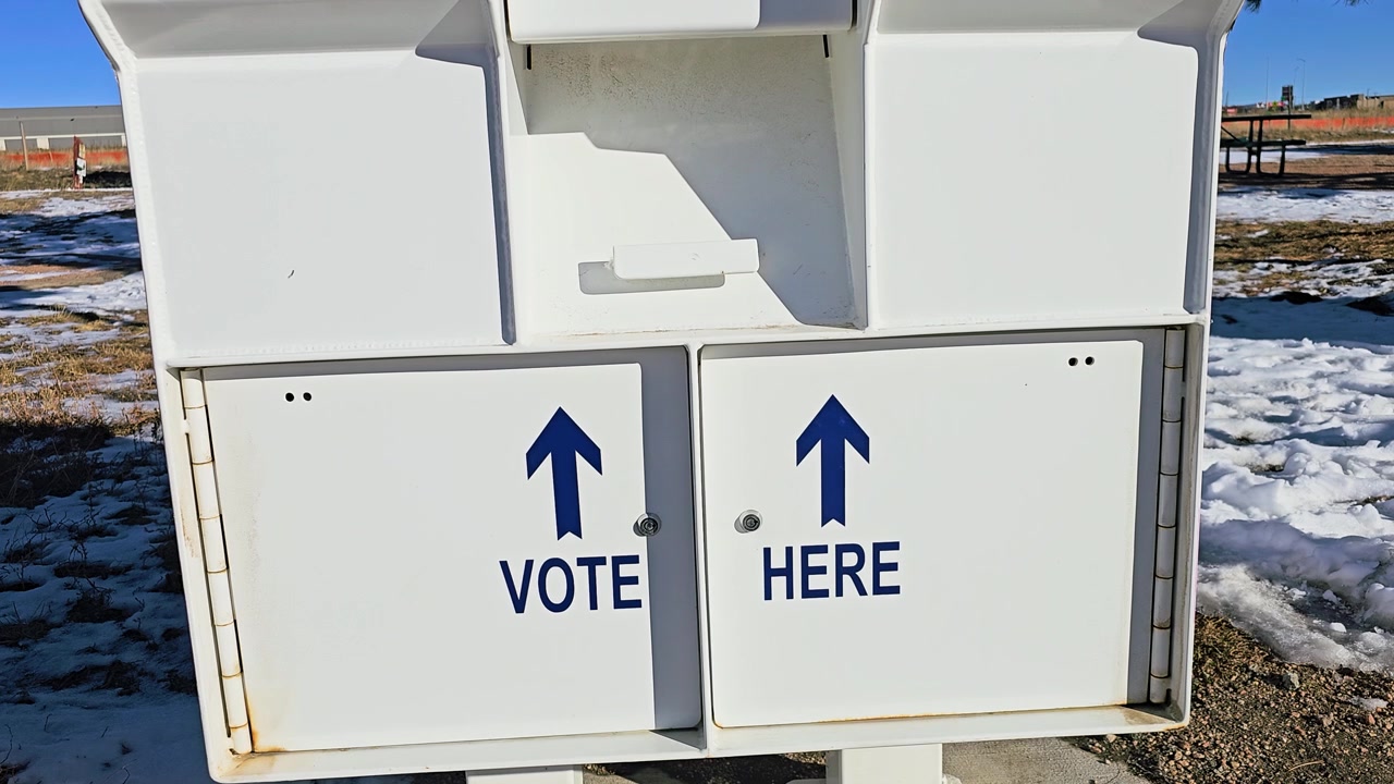 Secure election ballot box with Vote Here arrows, mail-in voting slots, surveillance warning, and Voting Closed sign standing outdoors beside snow-covered ground in bright winter daylight.