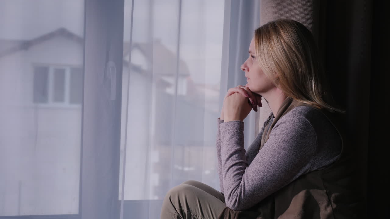 una mujer triste se sienta en el alféizar de la ventana mirando por la ventana a la casa