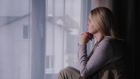 A-sad-woman-sits-on-the-window-sill-looking-out-the-window-at-the-house