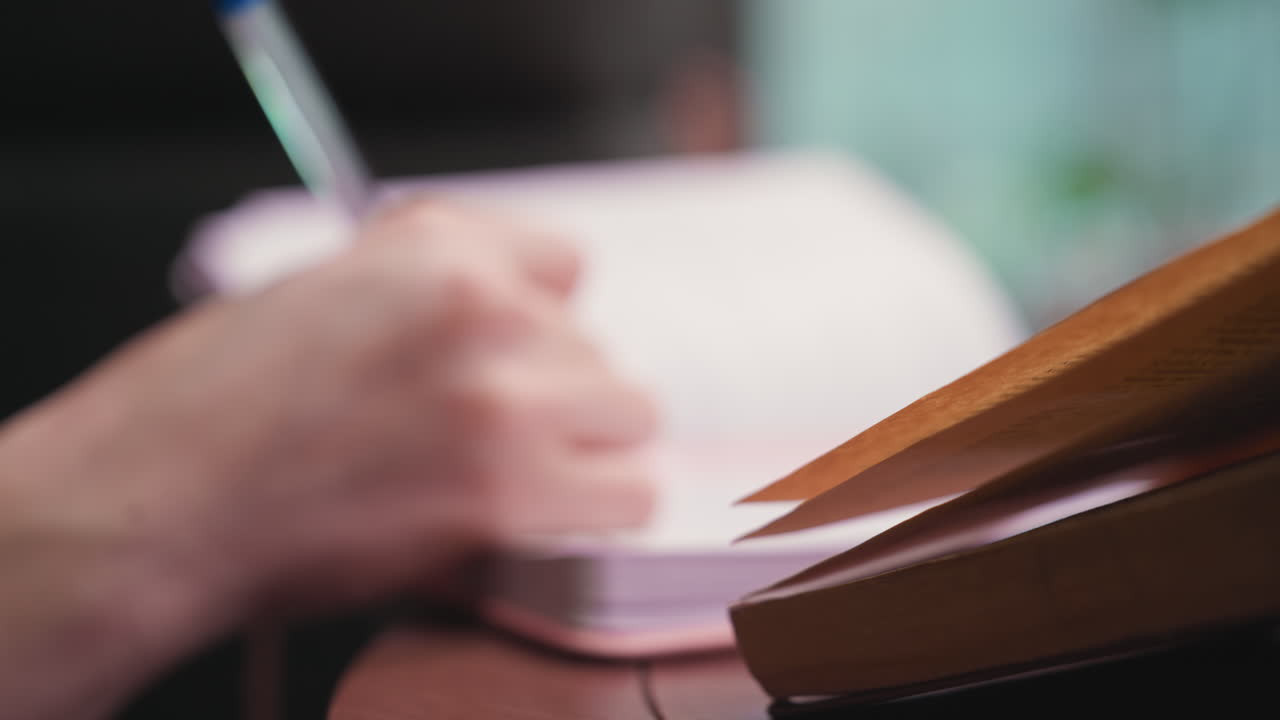 Blurred close up view of woman writing with blue pen in notebook beside open brown book on table, creating quiet reflective scene with shallow depth of field and soft ambient lighting