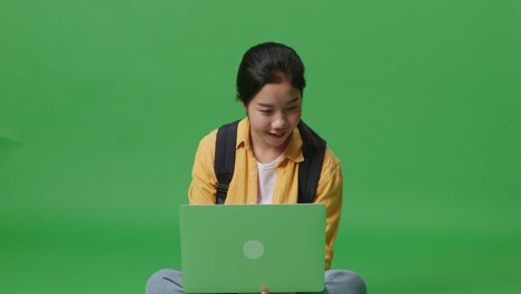 asian woman student with a backpack smiling and typing on a laptop while sitting in the green screen background studio