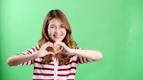 young asian woman with eyeglasses making heart symbol with hands in studio with chromakey