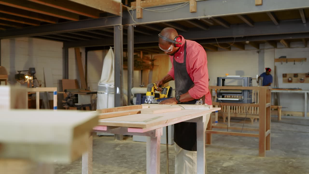 African American carpenter donning goggles, cutting boards with yellow jigsaw at workbench in shop