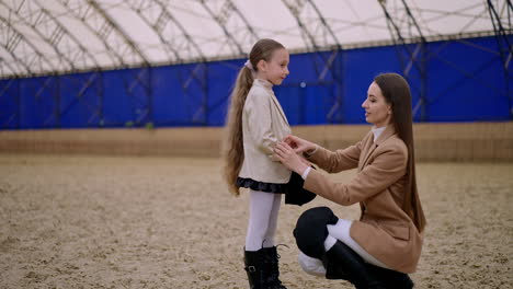 beautiful long-haired brunette woman and lovely girl at the manege. mother takes care of daughter's hair and clothes. blurred backdrop.