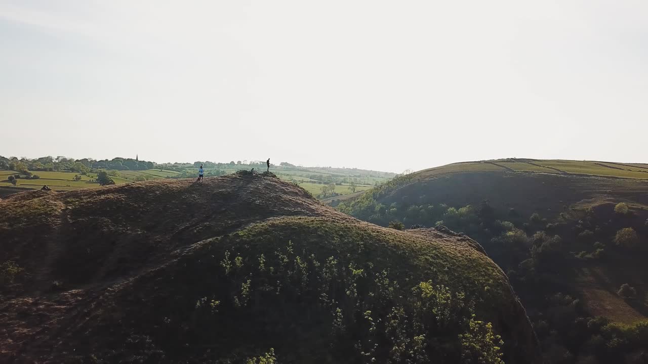 Aerial drone shot flying clockwise around people standing on top pf Mam Tor, Castleton, Peak District, England. Large hills and mountains, green fields and clear skies.