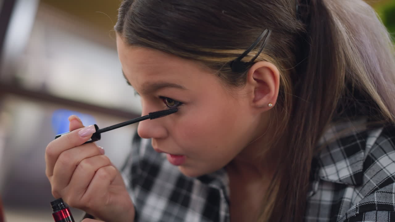 joven aplicando cuidadosamente máscara en el párpado inferior usando un pincel, centrándose en sus ojos frente a un espejo con un fondo borroso de un entorno público animado