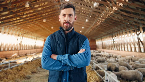 camera zooming on young caucasian man looking at camera with arms crossed in stable with sheep flock