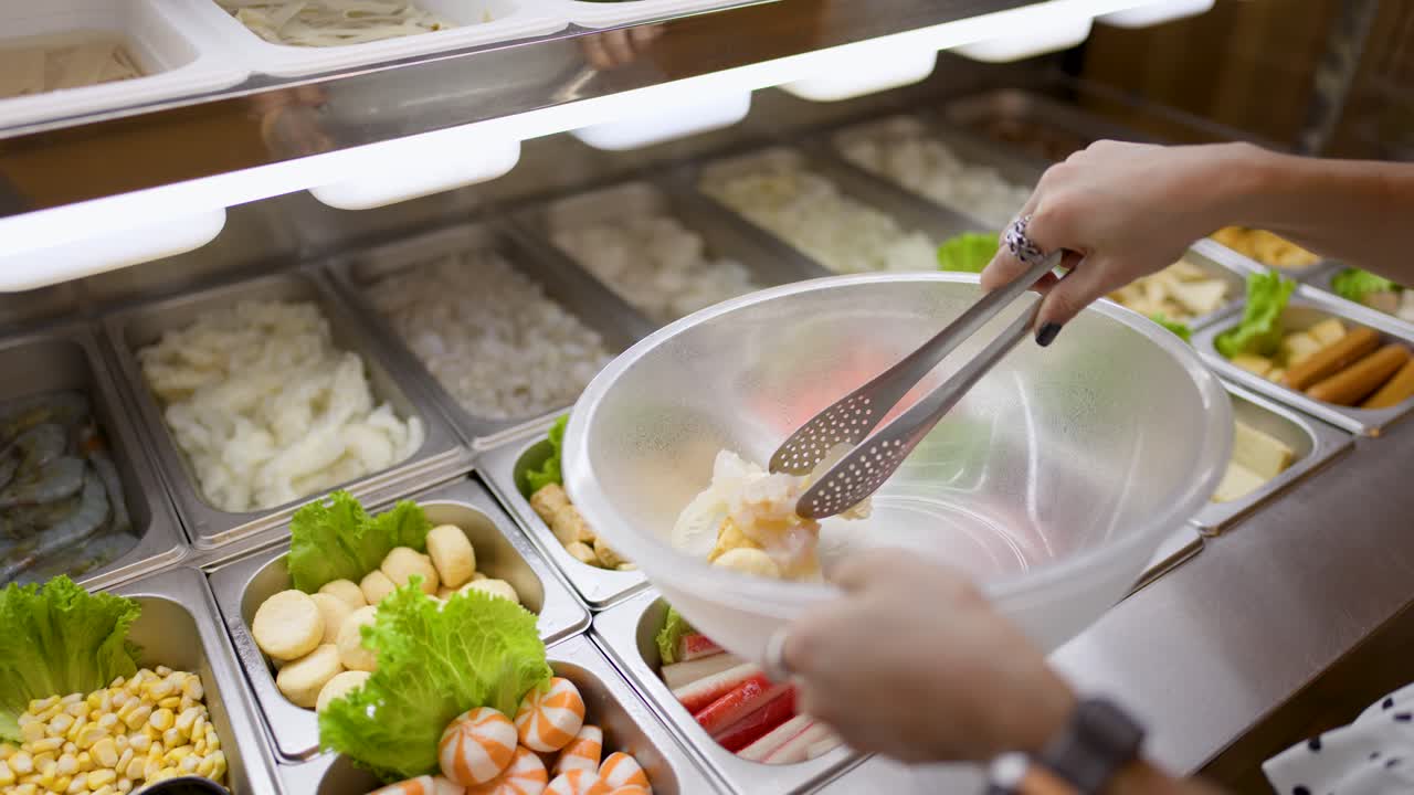 Person uses tongs to pick seafood and vegetables under bright lighting at buffet station