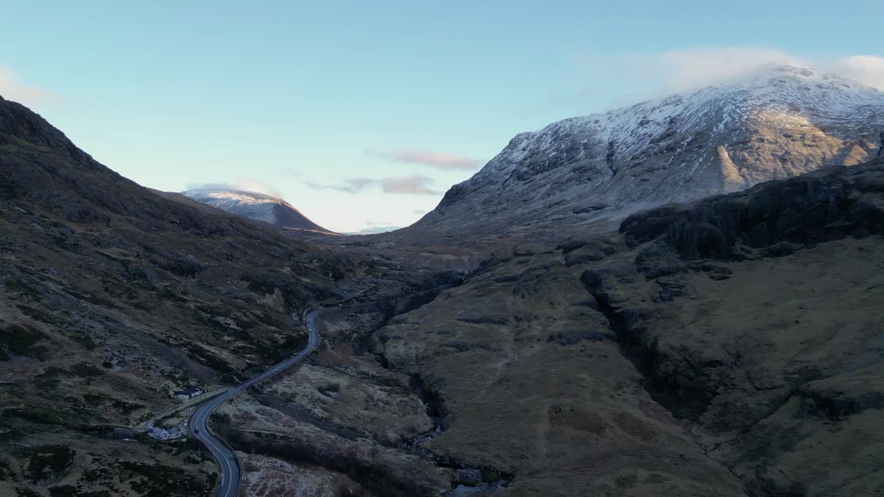Valle de Glencoe con una carretera sinuosa entre montañas escarpadas y nevadas al anochecer, vista aérea