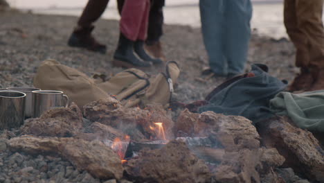 close up view of a bonfire with mugs and backpacks around it, in the background the legs of a group of teenagers on the seashore