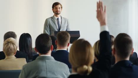 hombre de negocios caucásico con ropa formal hablando en una conferencia mientras una mujer levanta la mano para hacer una pregunta