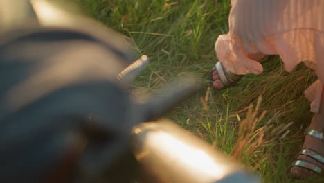 a close-up leg view of a woman wearing a flowing pink dress and sandals, gently dancing near a motorcycle in a grassy field