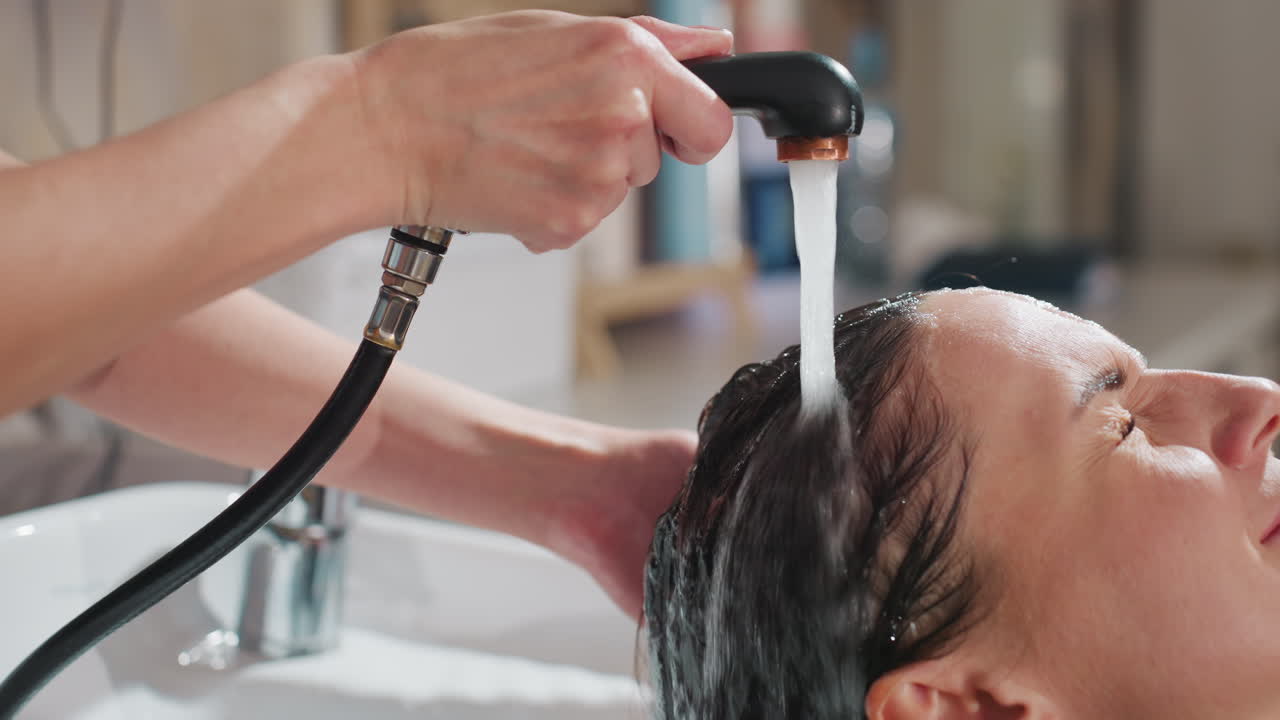 샴푸를 즐기며 싱크대에 기대어 있는 백인 여성, 스타일리스트가 휴대용 분무기로 따뜻한 물과 거품으로 머리를 헹구고, 목에는 수건을 받치고, 눈을 감고 편안하게 미소 짓고 있는 모습, 현대적인 미용실 세면대, 은은한 분위기