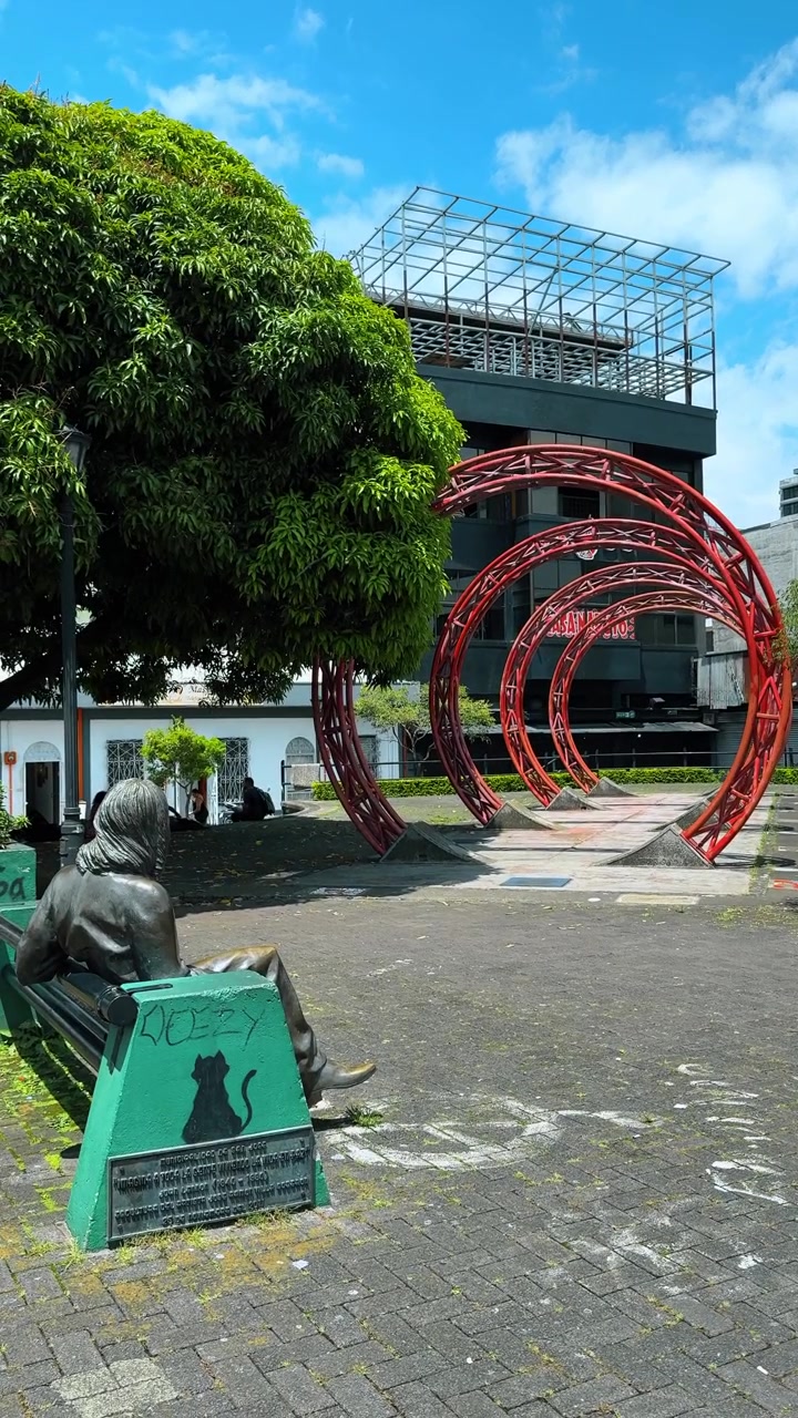 Escultura espiral roja en una plaza urbana, San José, Costa Rica. Anillos de metal junto a una estatua de bronce y un árbol verde bajo un cielo azul