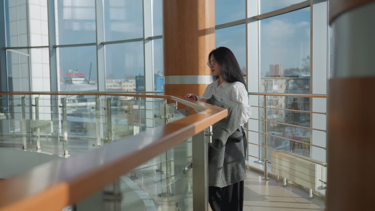 Young lady in white shirt holding checkered coat walks toward wooden railing inside bright modern mall, pauses to rest and gazes thoughtfully down and up through expansive glass windows