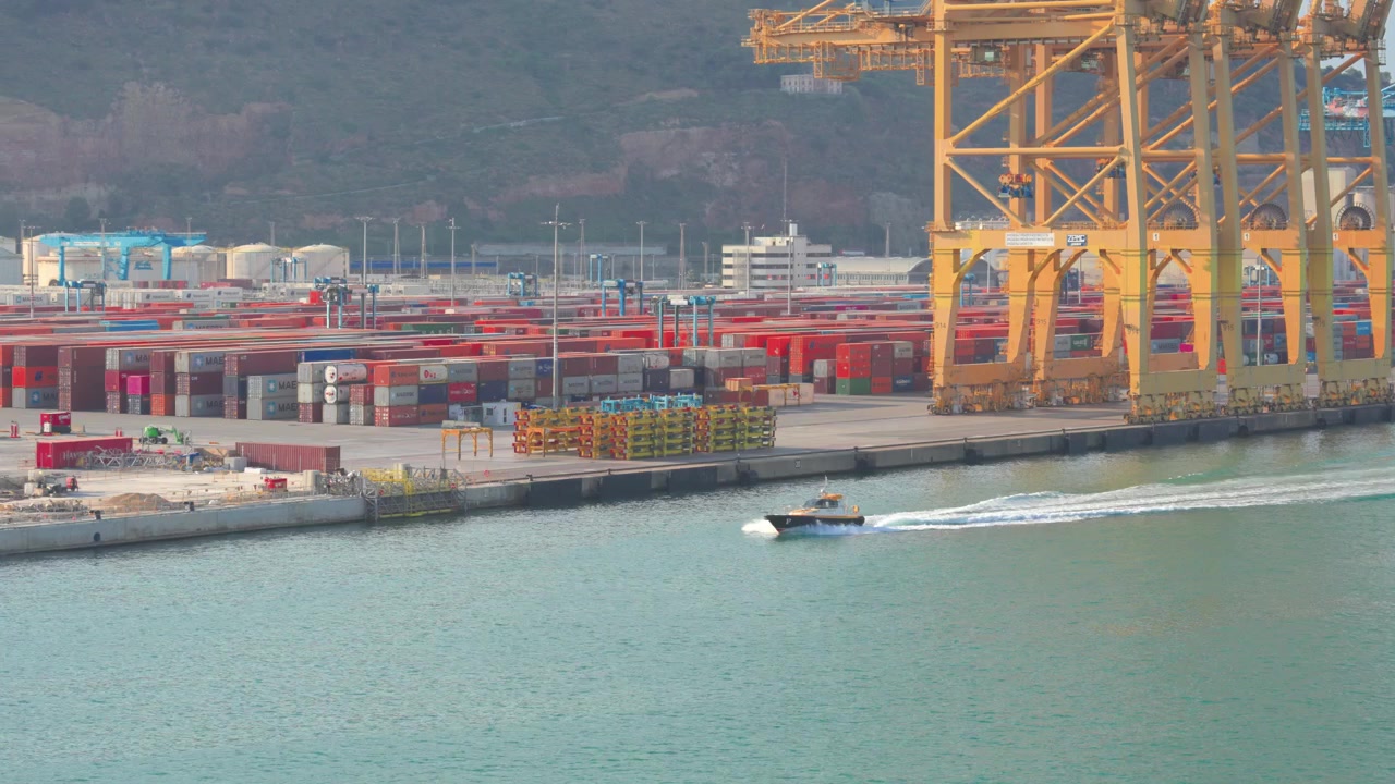 Small pilot boat navigating the waters of a busy industrial seaport, with towering gantry cranes and stacked shipping containers lining the commercial dock for global trade and freight
