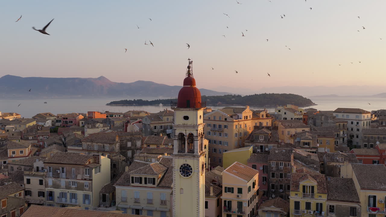 Drone circles around Saint Spyridon Greek orthodox church bell tower in Old town of Corfu with flock of swallows in flight around the town during sunrise
