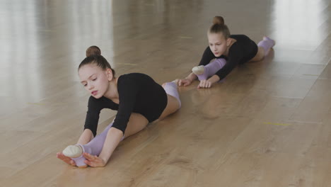 two gymnastic blonde girls with legs outstretched rehearsing a ballet move on the floor