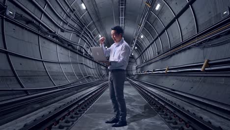 full body side view of an asian male professional worker standing with his laptop in underground subway tunnel, he raises his fist up with screaming goal after typed on the laptop