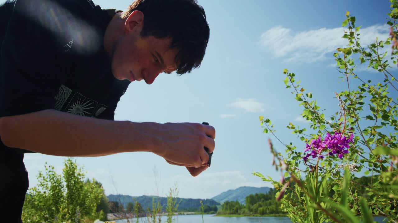 川のそばでスマートフォンで紫色の花の写真を撮る若い男性。