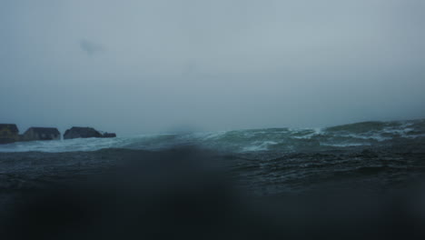 mist spray rises from top of wave, view from behind in water at surface of ocean on stormy grey day, slow motion