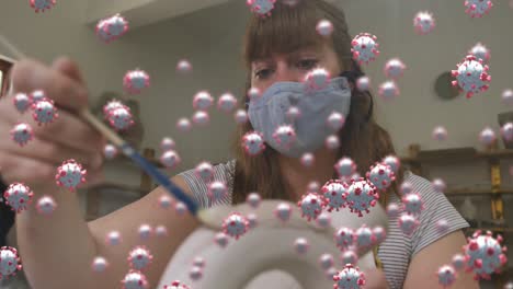 Animation-of-virus-cells-over-caucasian-woman-with-face-mask-painting-pottery