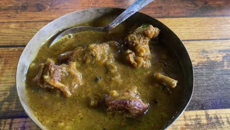 mutton curry is a dish that is served in a steel bowl on a wooden table and is also known as kosha mangsho, lamb curry, or goat curry