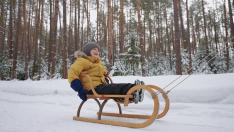 happy child boy is sitting in wooden sledge and riding over snow in winter forest family time