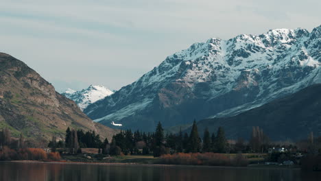 Airplane-Passing-By-On-Snowy-Mountain-With-Lake-Hayes-In-The-Foregrond-In-Queenstown,-New-Zealand