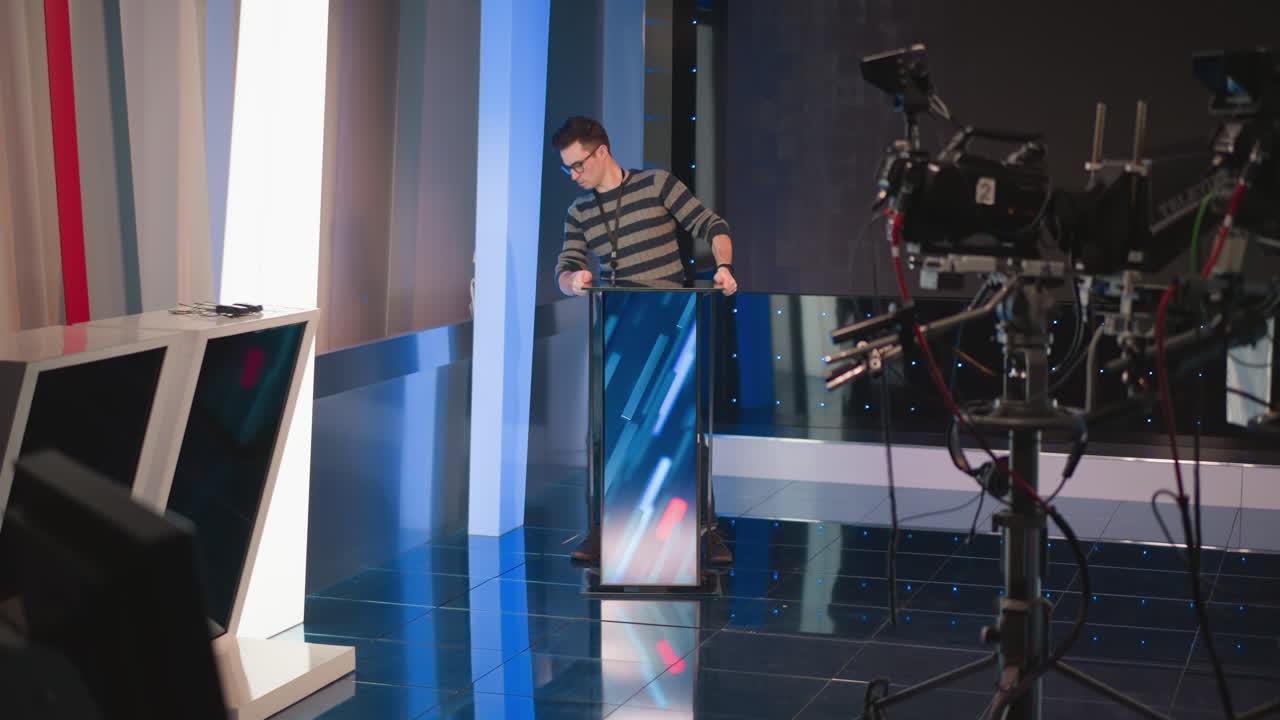 man adjusts digital display screen setup in modern video production studio, focused on screen configuration, with blue lighting, equipment, and camera on tripod in background, preparing stage
