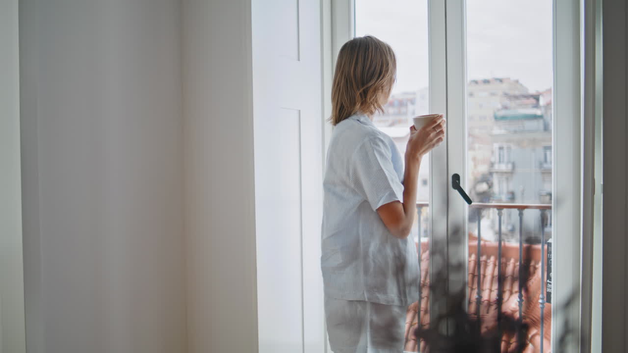 Woman enjoying coffee window in pajamas. Relaxed housewife looking city view