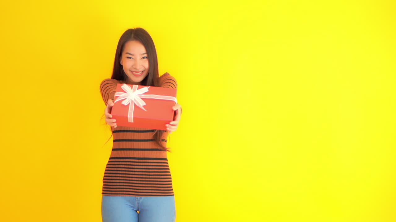 Static shot of a cute Asian lady holding a gift box while smiling in front of a yellow background