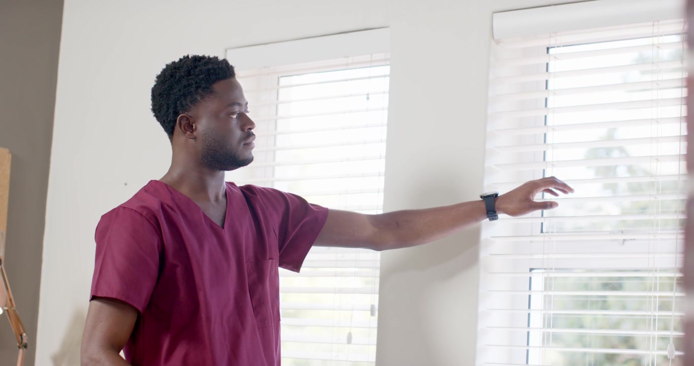African American man in burgundy scrubs checking watch, adjusting blinds to change light at clinic