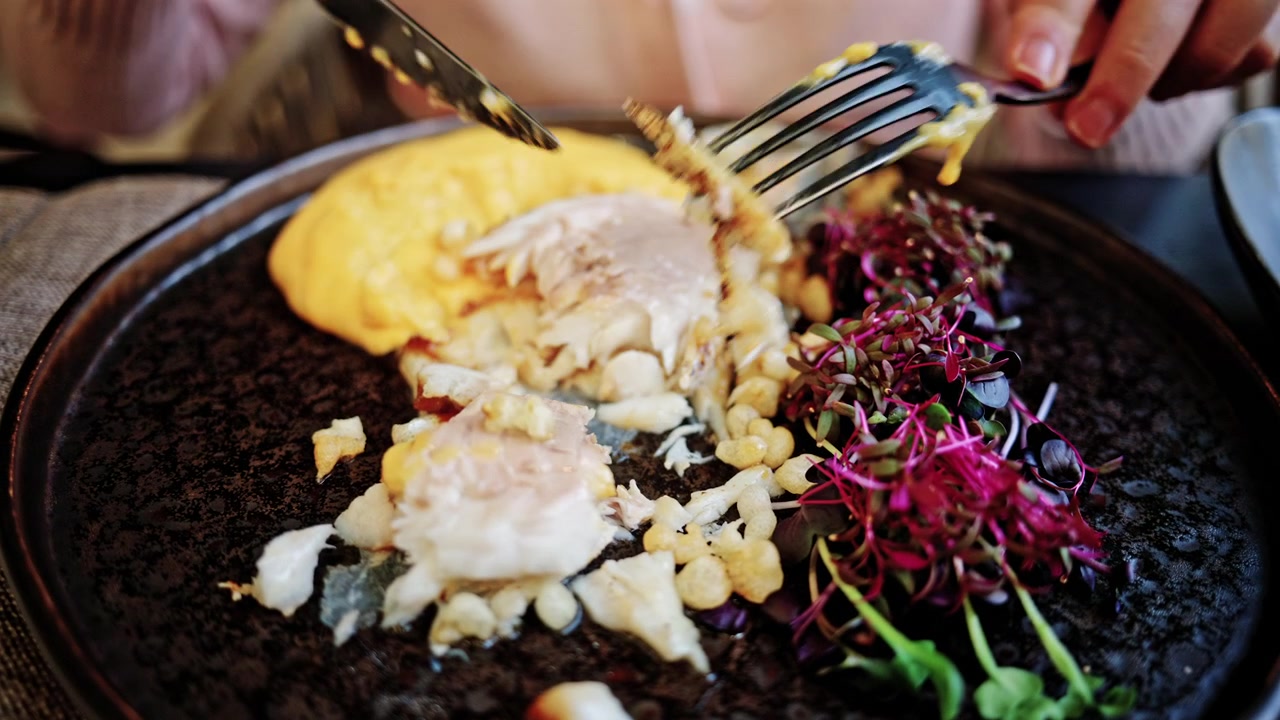 Primo piano di mani che usano forchetta e coltello per tagliare un filetto di pesce gourmet scottato con pelle croccante, impiattato con purè dorato e micro-verdure.