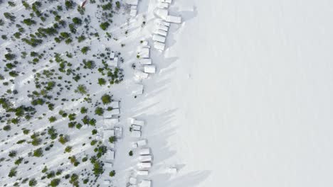 Flight-Over-Snowbound-Scandinavian-Landscape-Of-Forest,-Log-Cabins-And-Big-Lake