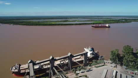 Buques-De-Carga-Y-Terminal-De-Granos-En-Un-Río-Fangoso,-Cielo-Despejado,-Vista-Aérea