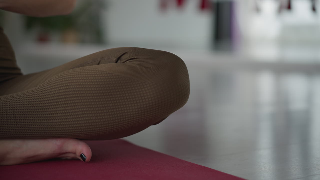 Upper body shot of wellness advocate seated cross legged on red yoga mat, lifting hands from sides to chest as she meditates peacefully in bright glass studio surrounded by hanging silk fabrics