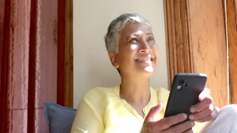 happy senior biracial woman sitting on couch and using smartphone at home, slow motion