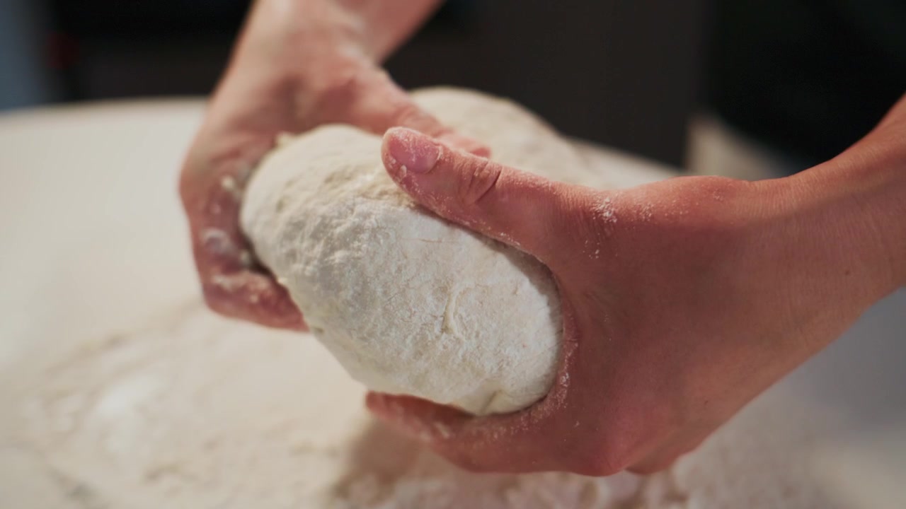 Mani tengono delicatamente una palla di impasto liscia e infarinata. La pelle della persona mostra residui di farina, suggerendo un'impastatura attiva. La superficie sottostante è coperta da farina bianca fine.