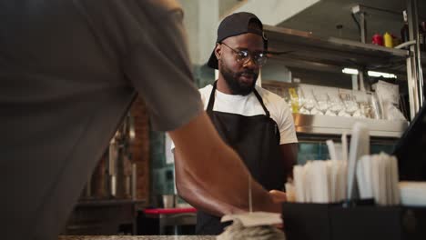 a black person in a cap works in a doner market. a man with glasses takes an order and makes a check at a doner market