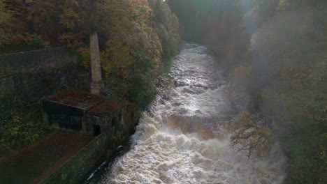 imágenes de drones en cámara lenta sobre un río que fluye rápidamente antes de descender hacia una cascada rodeada de edificios y un bosque de árboles de hoja ancha y coníferas