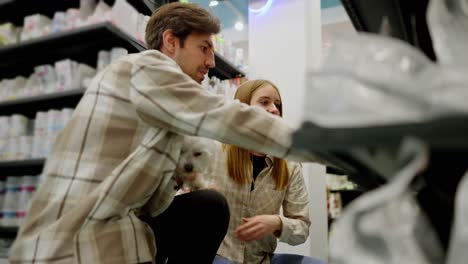 a brunette guy in a light checkered shirt is talking to a blonde girl and her white dog about purchasing items in a pet store