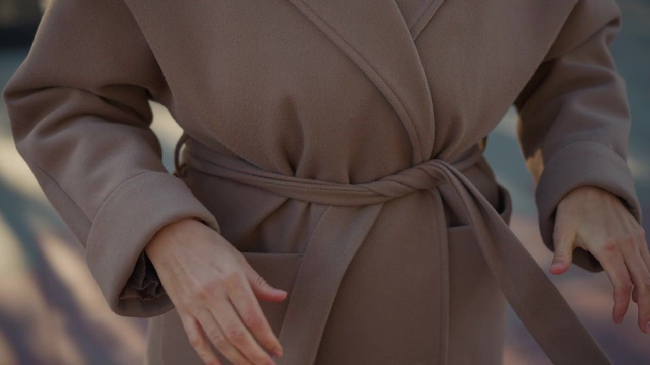 Primo piano di una donna che allaccia la cintura del suo cappotto marrone, concentrandosi sulle mani e sulla manipolazione del tessuto. L'azione mostra un'attenta regolazione per la vestibilità, con una morbida illuminazione che evidenzia i dettagli della texture.