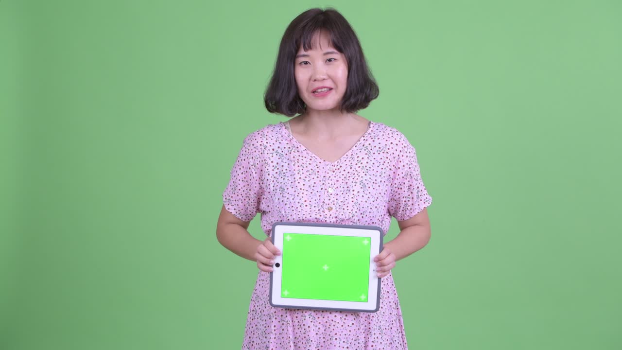 Studio shot of beautiful pregnant Asian woman holding digital tablet with copy space against green studio background